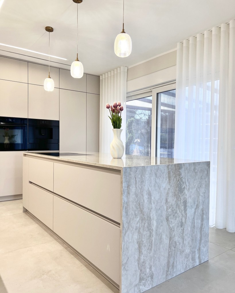 Moderne Kücheninsel aus hellen Materialien mit eleganter Steinoberfläche, minimalistischen Schränken und frischen Blumen in einer weißen Vase, umgeben von großen Fenstern und sanften Vorhängen.