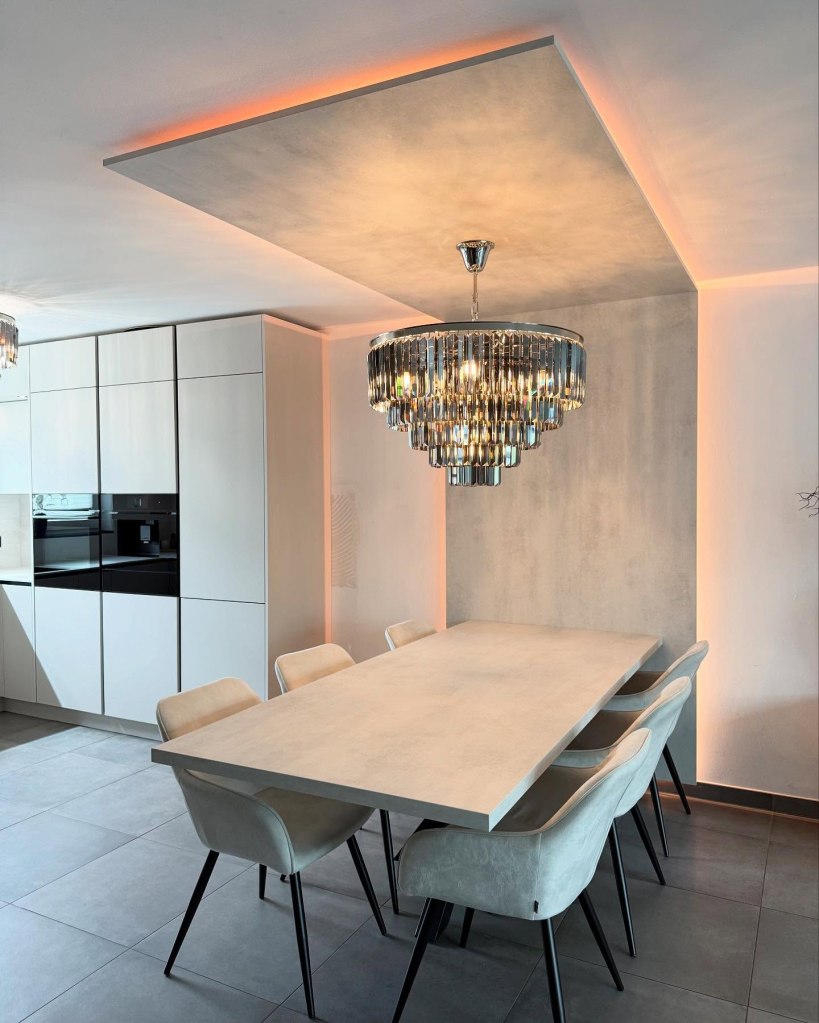 Ein modernes Esszimmer mit einem großen, rechteckigen Tisch und eleganten Stühlen. Eine auffällige Pendeleuchte aus Kristall hängt von der Decke, während indirektes Licht die Wände sanft beleuchtet. Küchenschränke im Hintergrund.